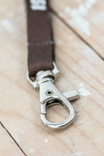 Lade das Bild in den Galerie-Viewer, Auf dem Bild sieht man einen Holzboden. Auf diesem liegt ein kleiner Karabiner mit braunem Band.
