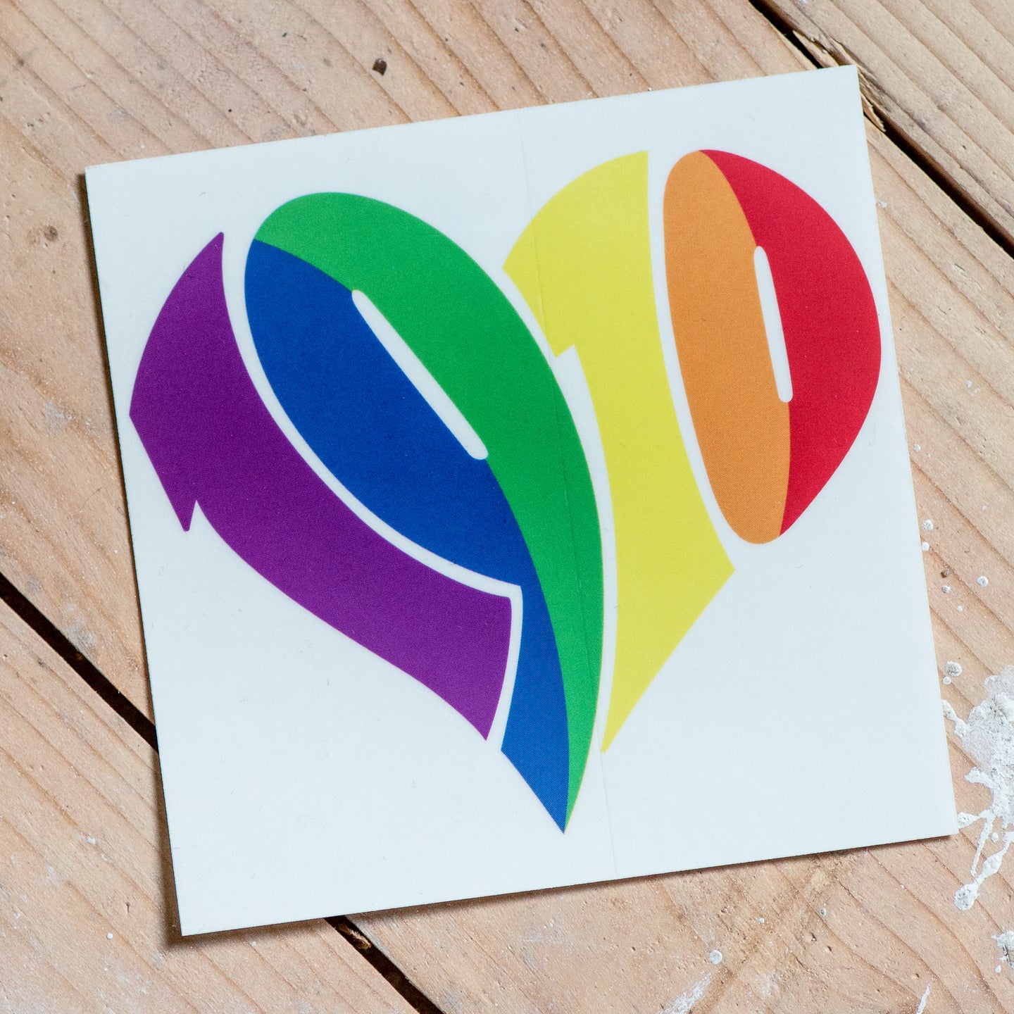 Auf dem Foto sieht man einen Holzfußboden. Auf diesem liegt ein quadratischer weißer Sticker mit großem 1910 Herz in regenbogenfarben.