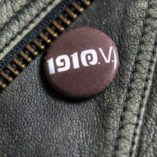 Lade das Bild in den Galerie-Viewer, Auf dem Bild erkennt man eine schwarze Lederjacke. An dieser hängt ein brauner Button. Auf diesem befindet sich das weiße Logo 1910 eV.
