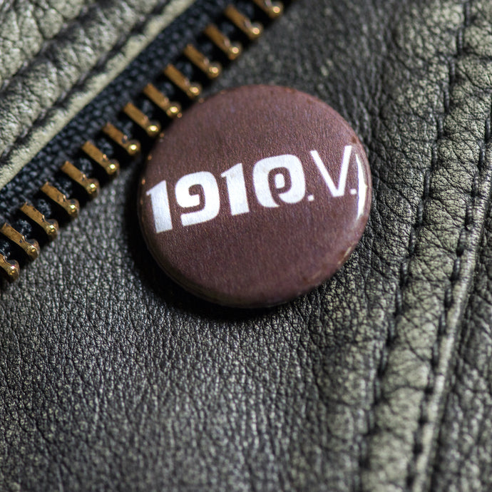 Auf dem Bild erkennt man eine schwarze Lederjacke. An dieser hängt ein brauner Button. Auf diesem befindet sich das weiße Logo 1910 eV.