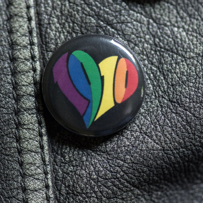 Auf dem Bild sieht man einen schwarzen Button mit einem großen regenbogenfarbenen 1910 Herz darauf. Der Button befindet sich auf einer schwarzen Lederjacke.